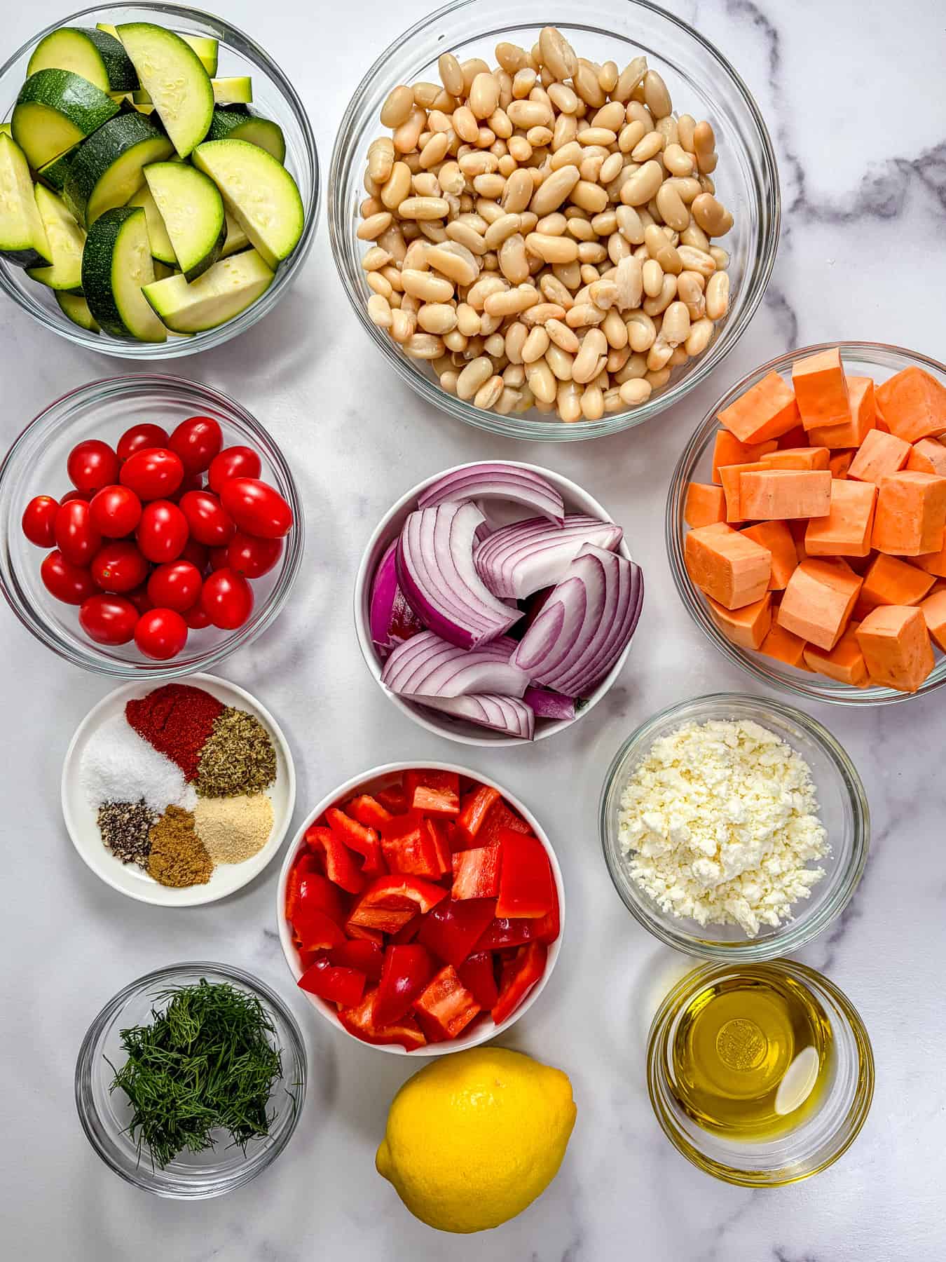 White beans, zucchini, squash, red onion, cherry tomatoes, feta, olive oil, and herbs in glass bowls on a marble background.